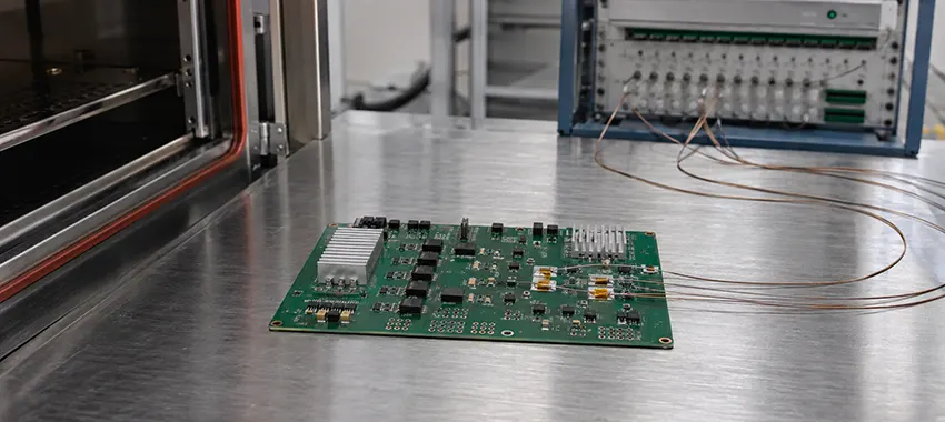 Multilayer PCB with thermocouples taped to the board on a lab bench beside a thermal test chamber